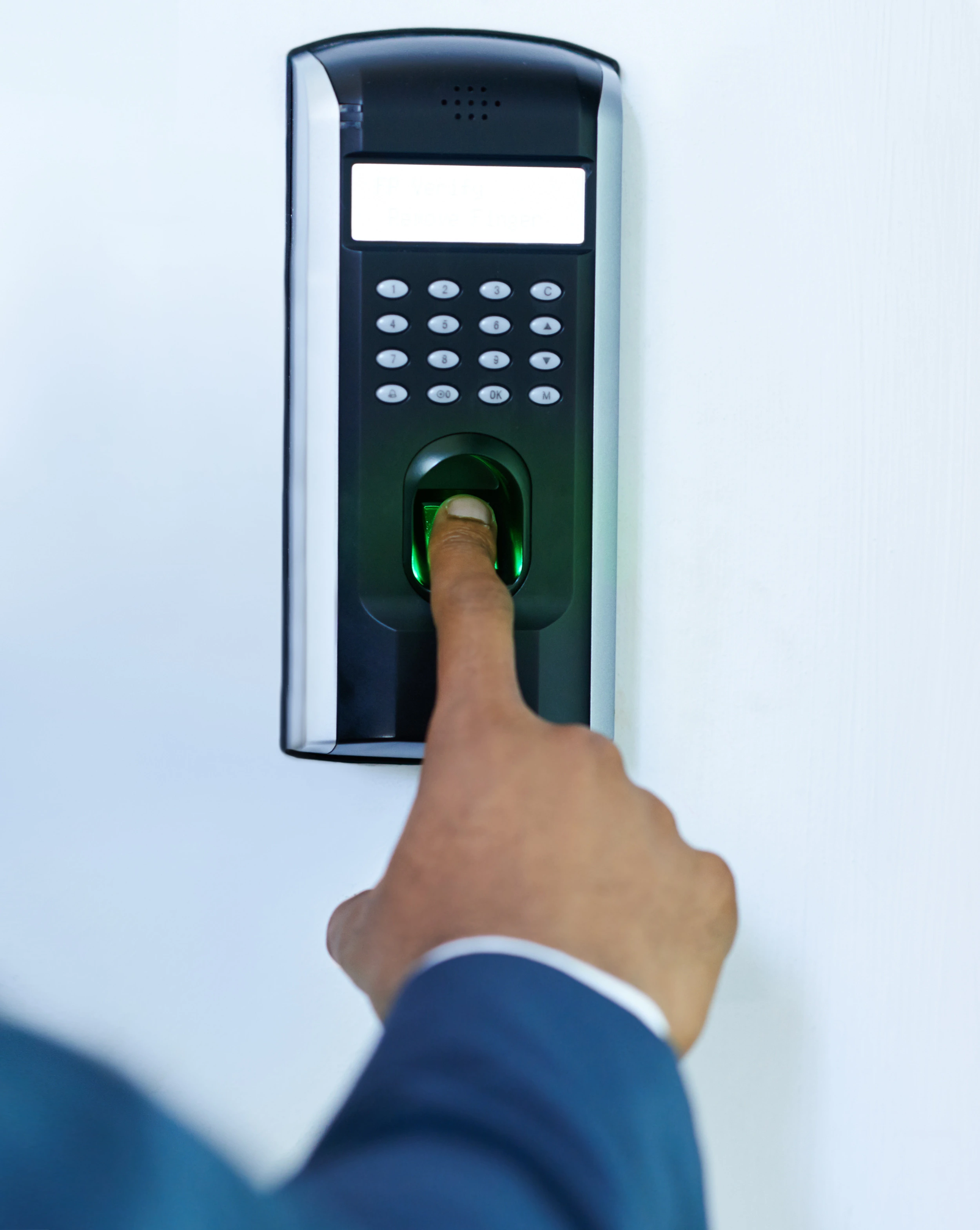 Fingerprint scanner with a green light on a wall. Blurred background shows two people standing in a bright hallway with bulletin boards.