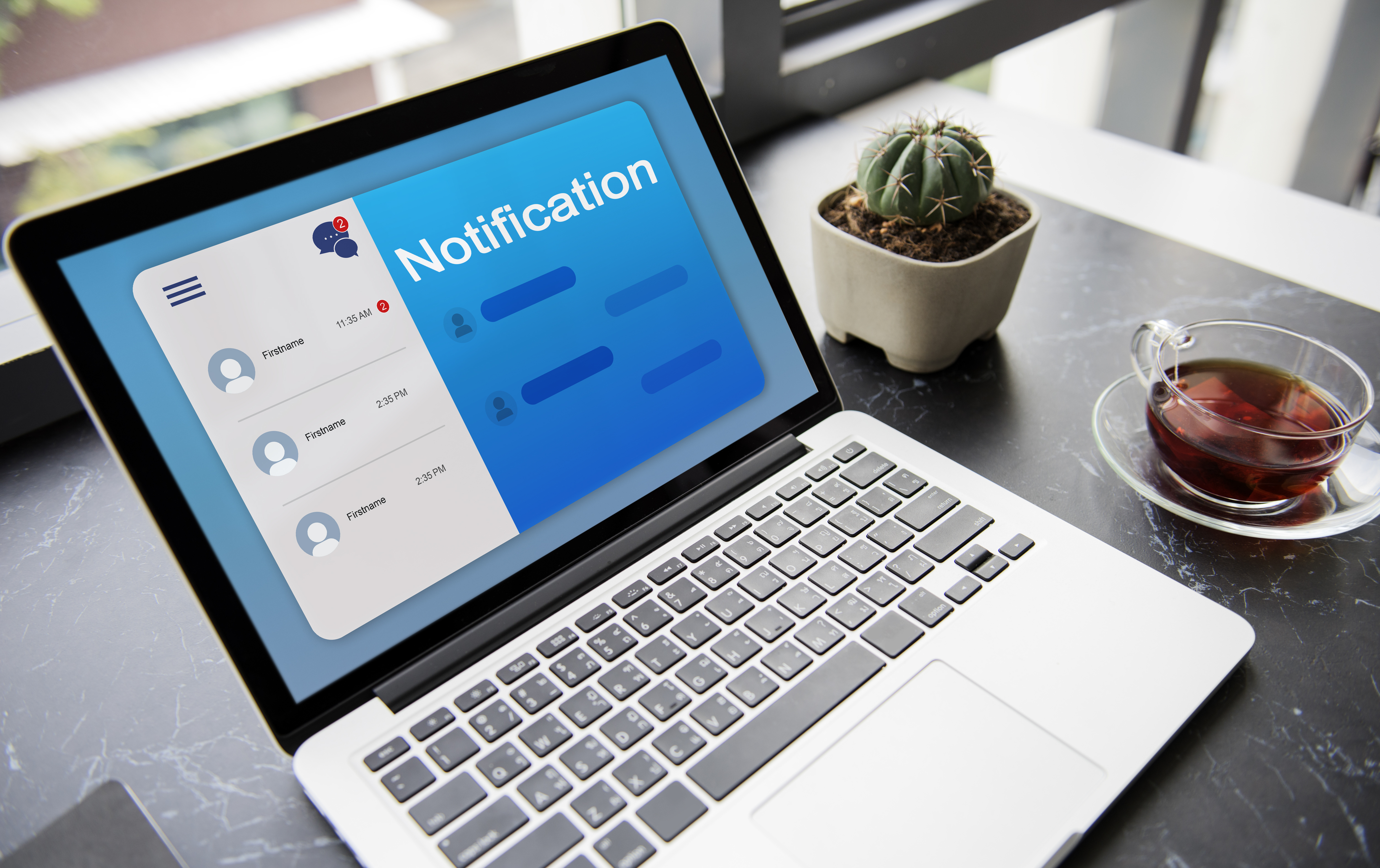 Laptop on a black marble desk shows a blue notification screen. A cactus and a glass cup of tea are nearby, creating a calm setting.