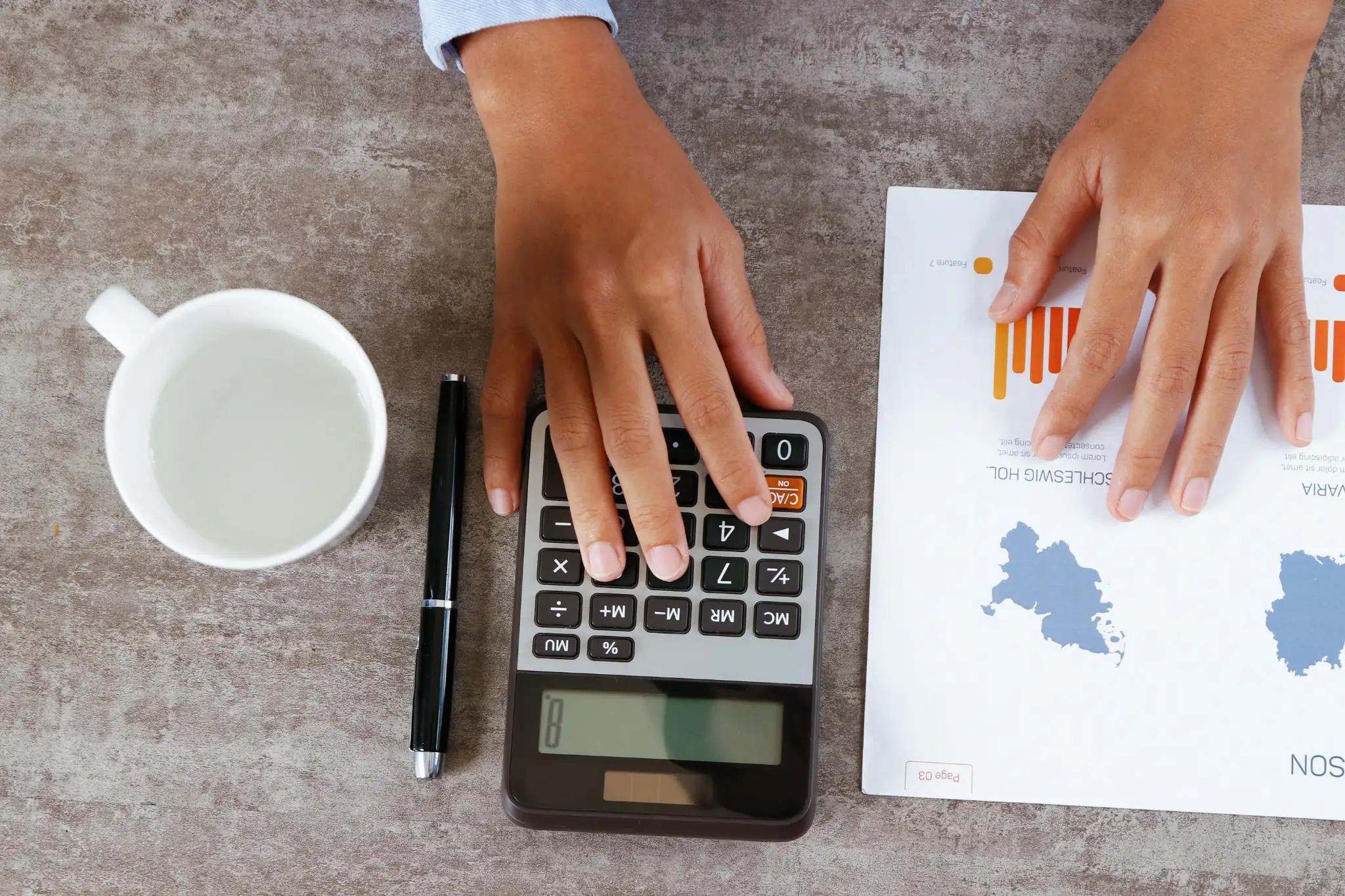 Hands using a calculator and holding a report with graphs, beside a cup of water and a pen on a gray desk.