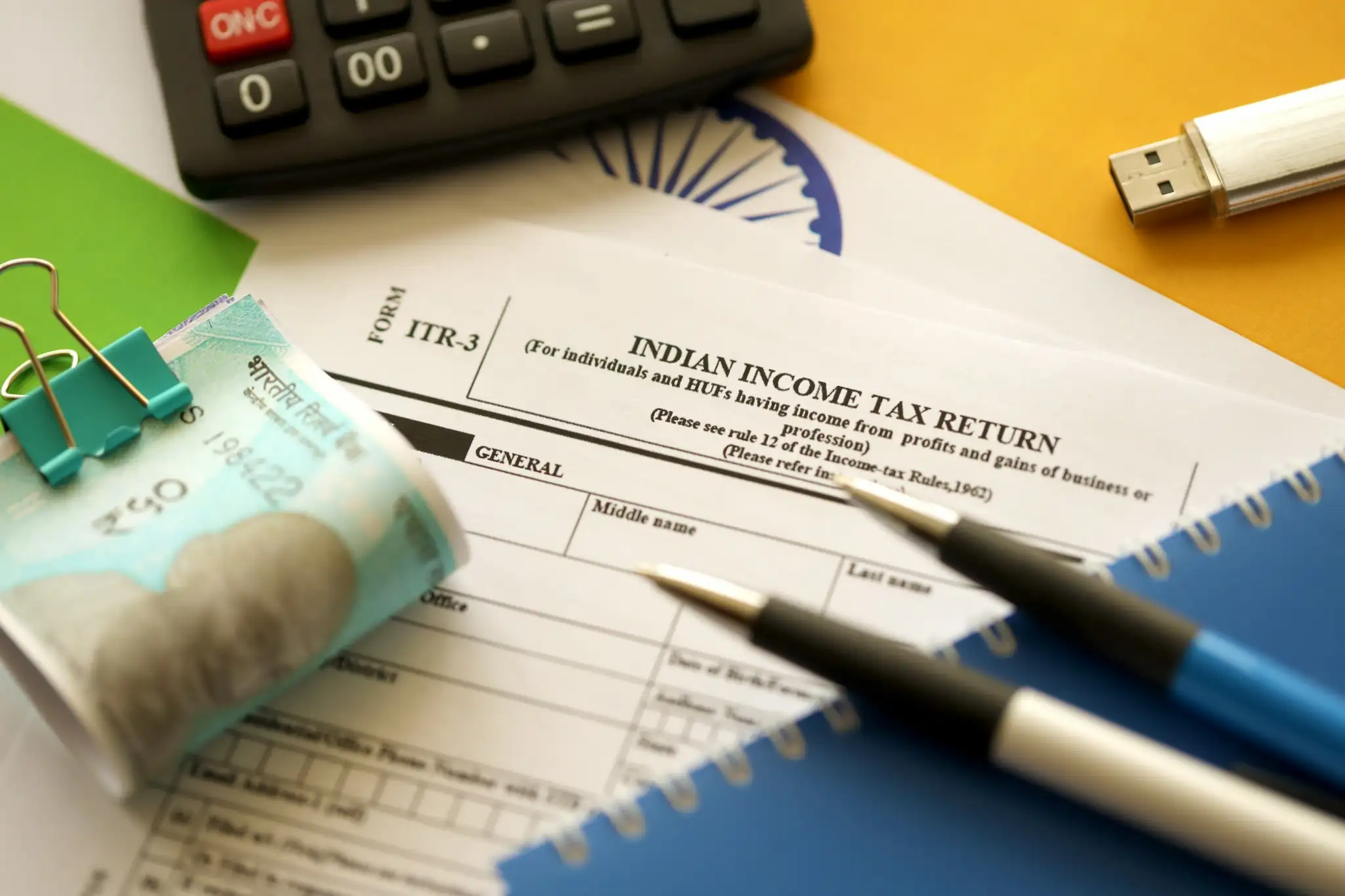 Indian Income Tax Return form on a desk with pens, a calculator, currency notes, a USB drive, and a green binder clip.