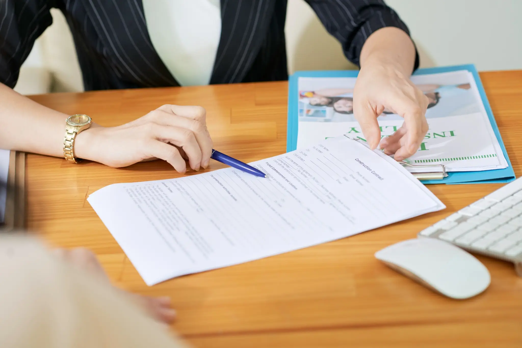 Hands reviewing a contract with a pen on a wooden desk. A blue folder and keyboard are visible. Business setting.